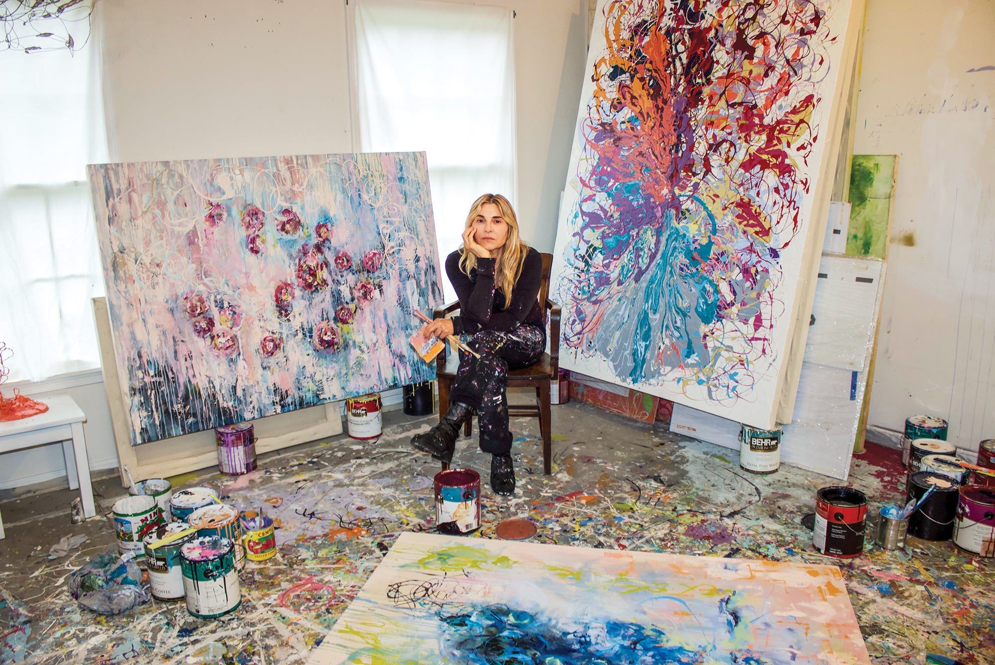 A female artist sits in her studio surrounded by her paintings and artwork.