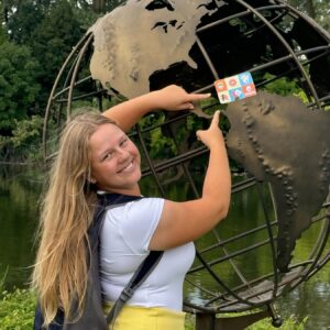 a student smiles at the camera. she wears a backpack. she is pointing at a Grubhub gift card that sits on top a statue of a globe.