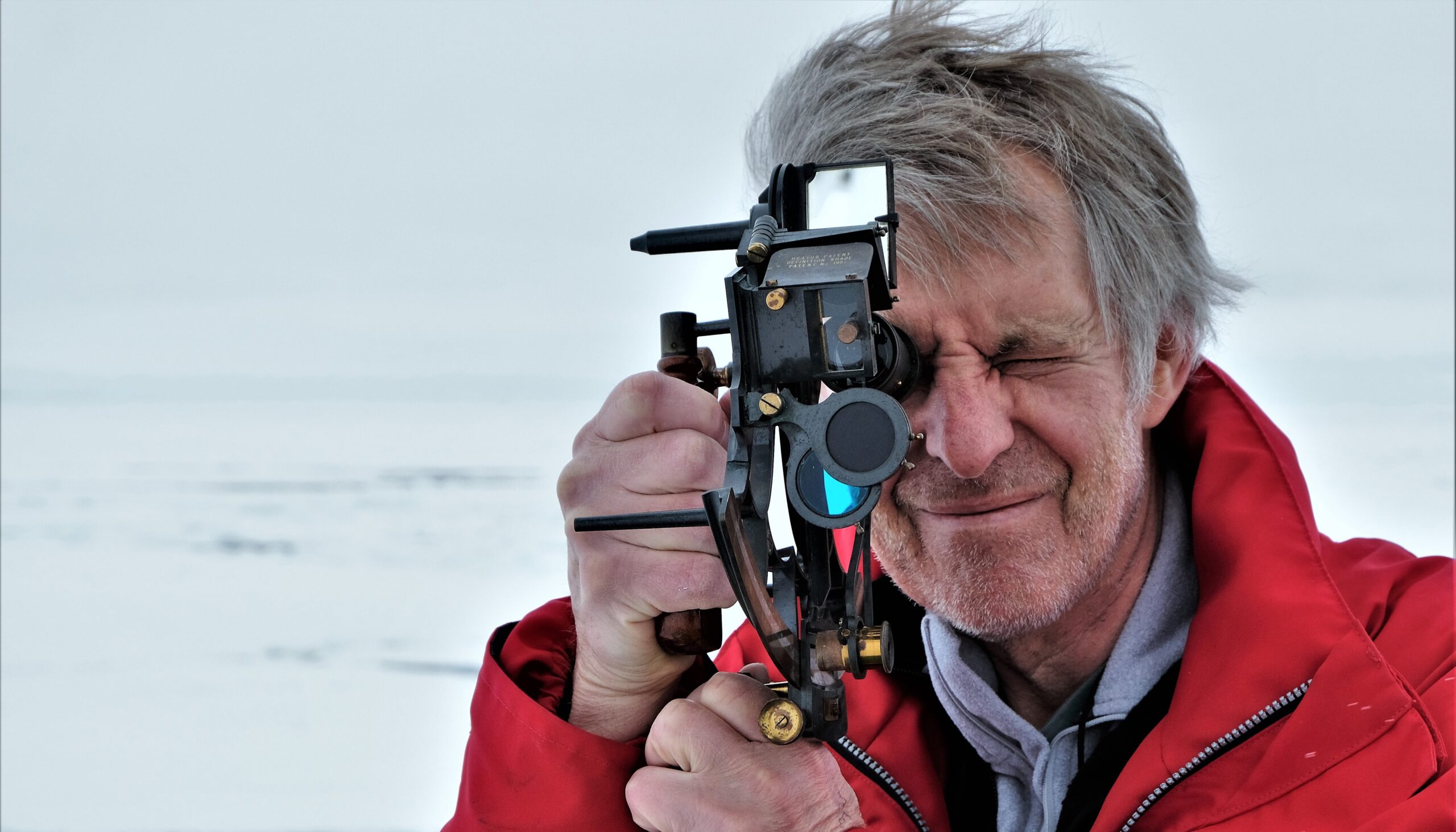 A man takes an angle on the sun with a sextant. He wears a red parka coat and stands on the ice.