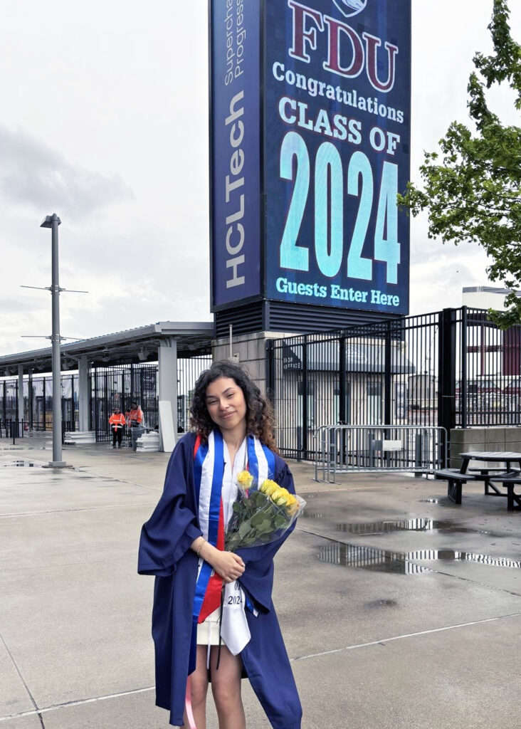 A young woman in a graduation robe pose for a photo outside of a stadium.