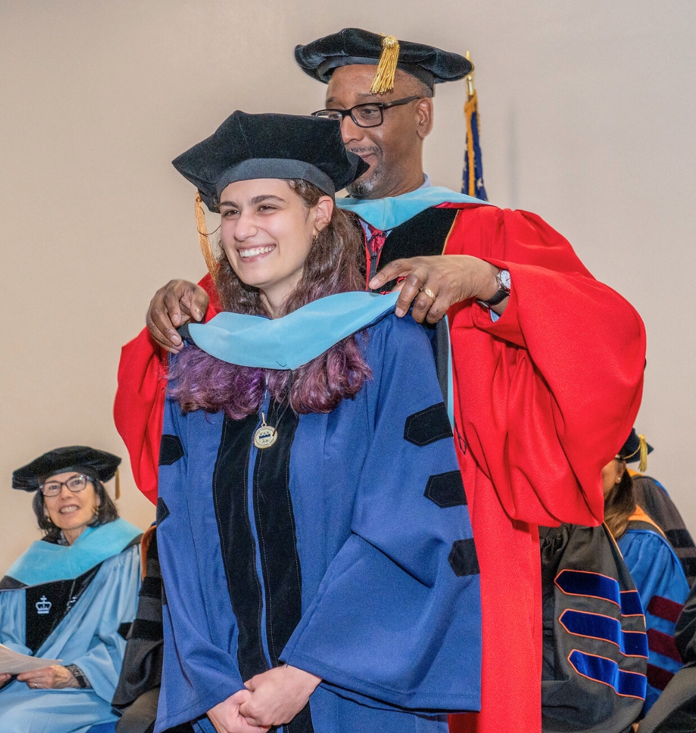 a faculty member puts a doctoral hood around a graduate. they both smile.