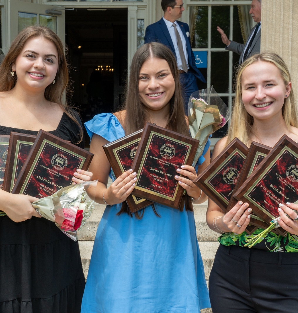 three girls smiling at the camera. they all hold flowers and multiple awards.