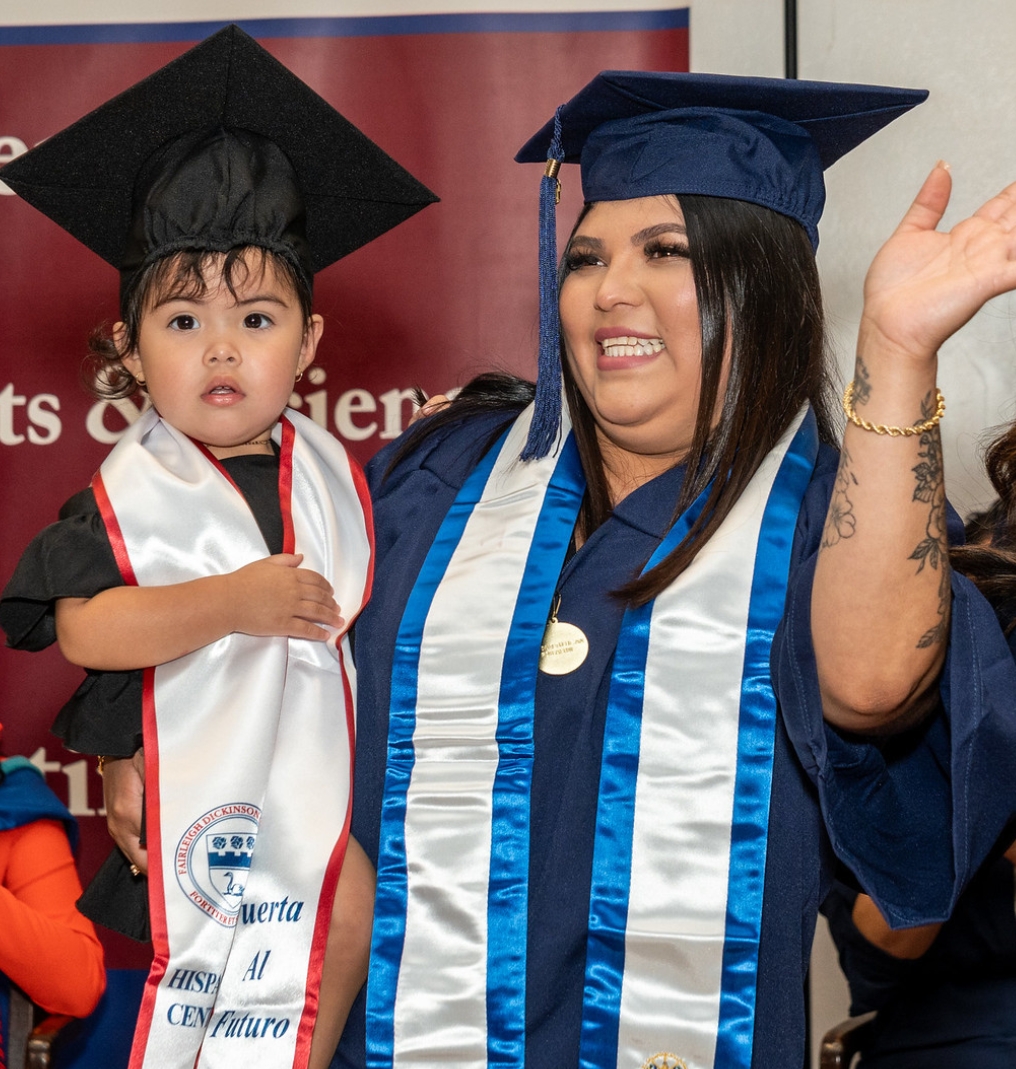 a graduate holding a child. they both wear a cap and gown. the graduate waves at someone off camera.