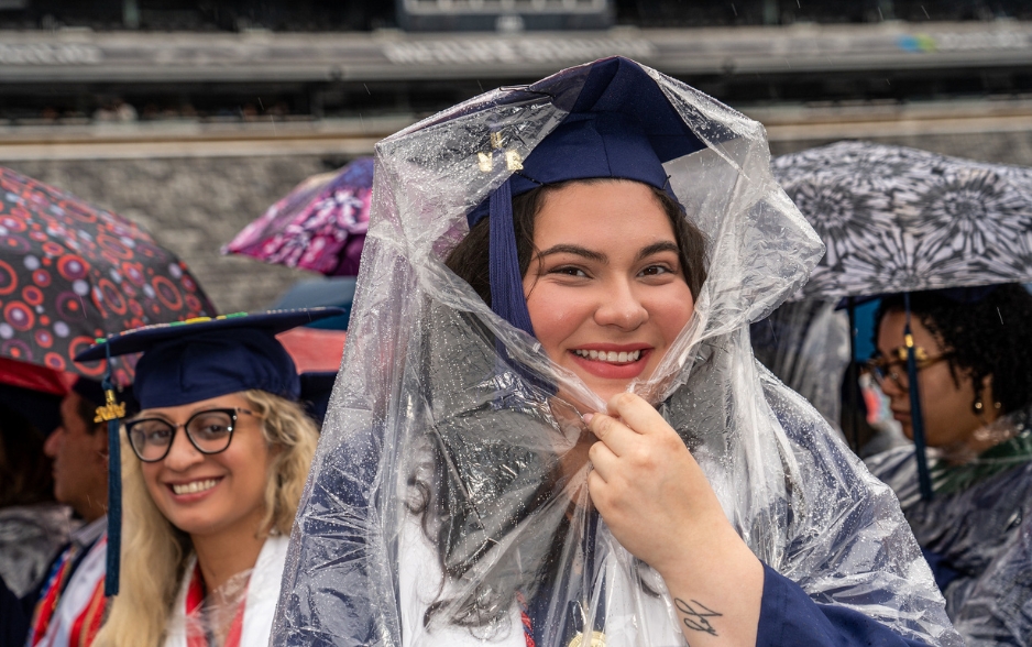 a graduate smiling for the camera. she holds a rain cape over her head.