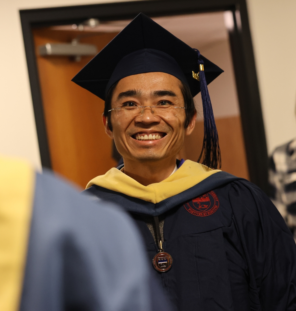 graduate smiling at the camera. he wears a cap and gown.