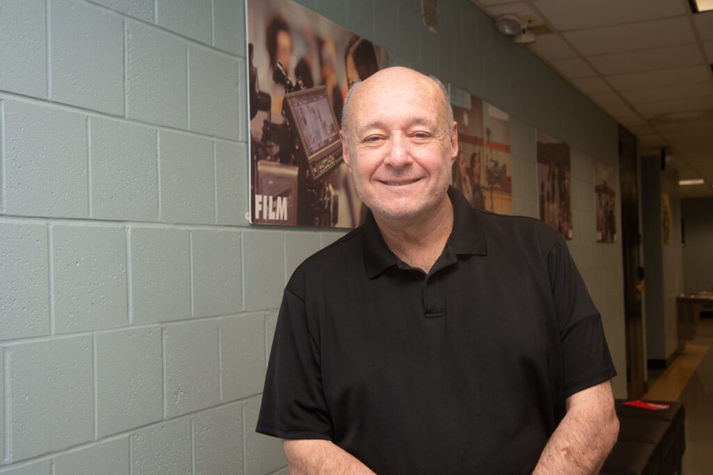 a person standing posing for the camera. there is a poster of a camera behind him.