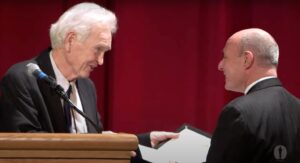 two men smile at each other in front of a red stage curtain. one is at a podium and hands the other an award.