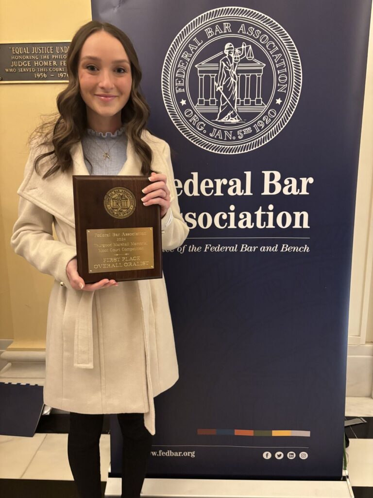 a student stands in front of a banner that reads "federal bar association." she holds up an award plaque.