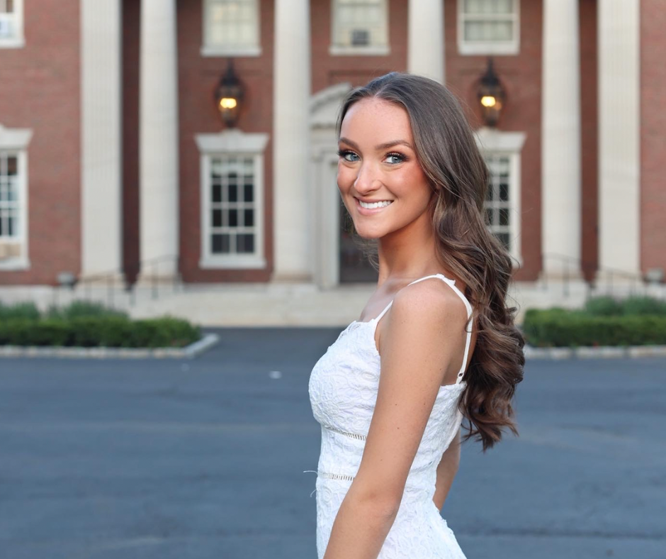 a student stands in front of a large estate mansion. she wears a white dress.