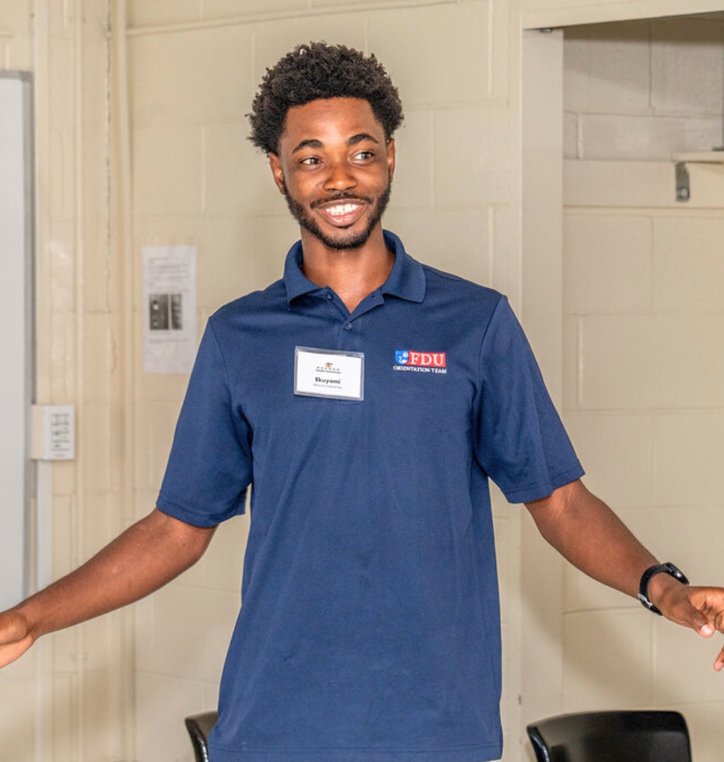 a student smiles at something out of frame. he wears an FDU-branded polo shirt and name tag. he demonstrates something with his arms outstretched.