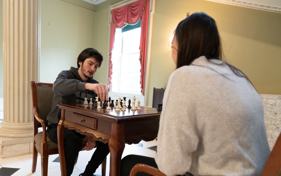 a student sits and plays chess. he wears an FDU hoodie. the back of another student playing against him can be seen in the edge of the photo.