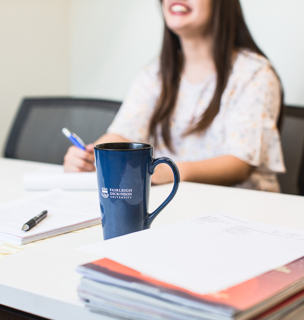 A desk cluttered with papers and notebooks. An FDU-branded mug sits on the desk. A woman is in the background.