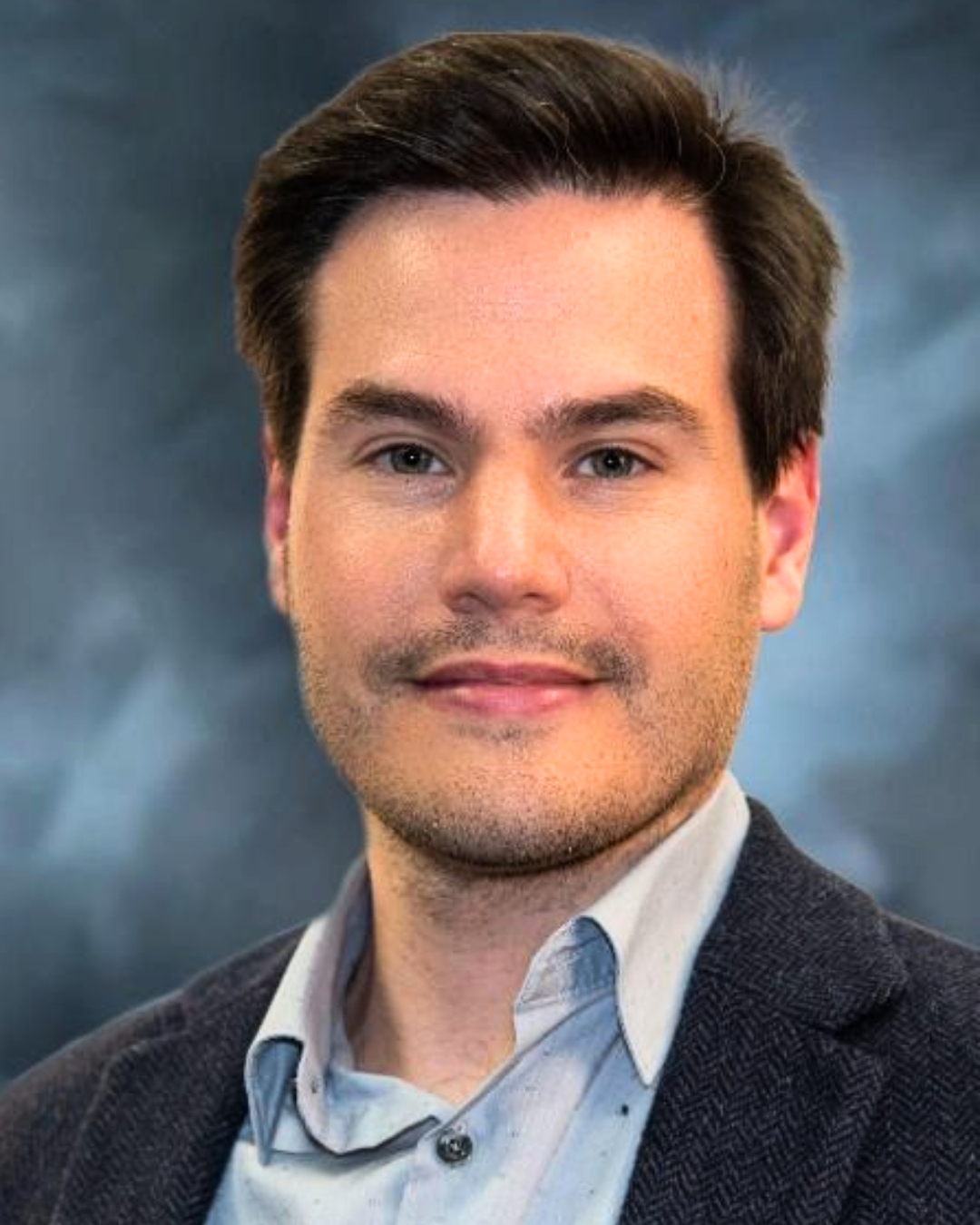 portrait of a man against a gray backdrop. he wears a blazer.