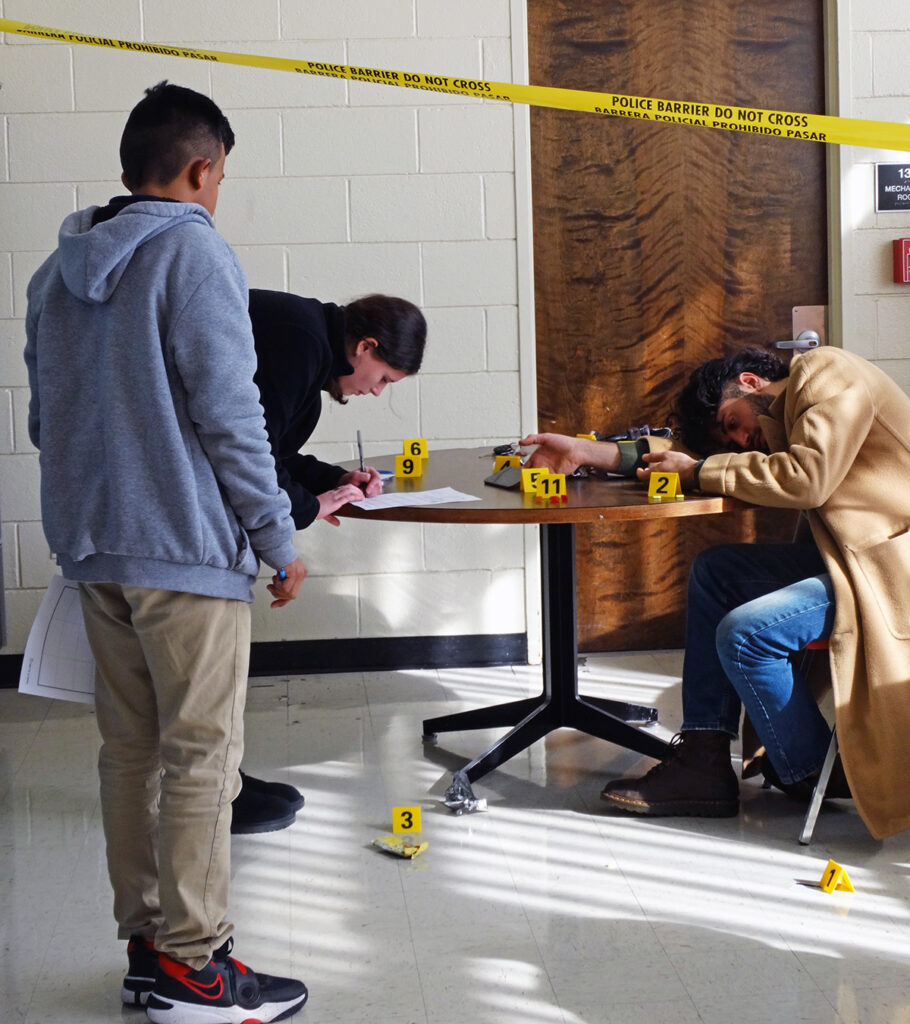 A faux crime scene is set up with police tape and evidence markers. A college student plays a corpse.