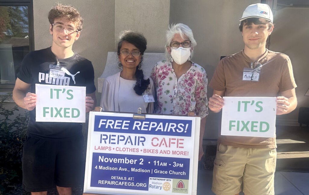 a group smiles for the camera. they hold signs that read "it's fixed," and "repair cafe."