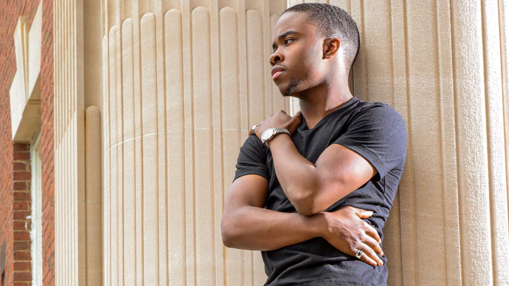 A young man leans back against the exterior pillar of a large brick building.