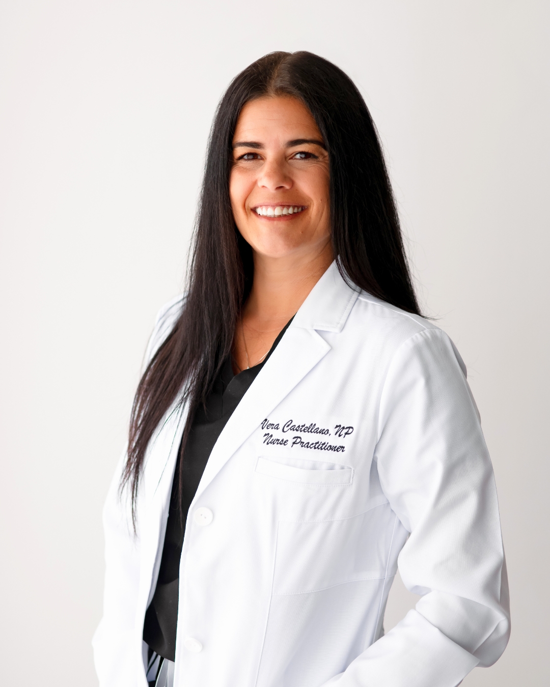 portrait of a woman against a wall. she wears a white lab coat with her name embroidered on it.