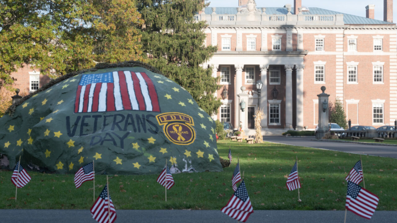 a large rock in front of the Hennessy Hall mansion. an American flag and gold stars are painted on the rock. the rock reads "Veterans Day." American flags are standing in the grass surrounding the rock.