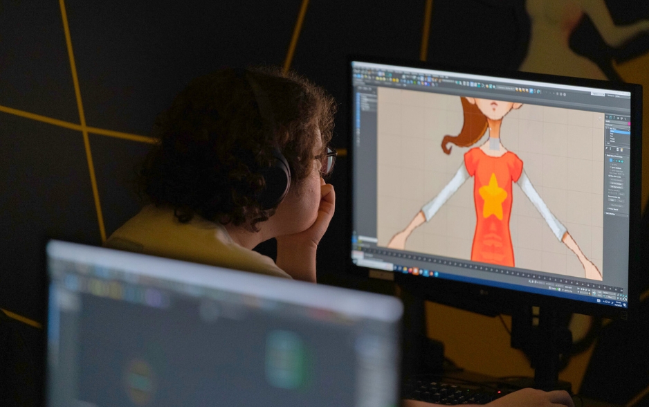 a student looks at a desktop computer. he wears headphones. the screen shows an unfinished animation of a girl with a ponytail.