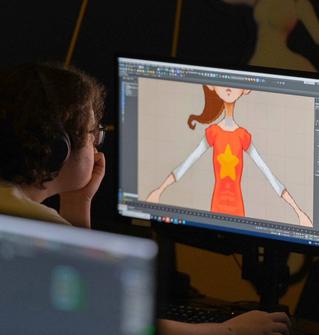 a student looks at a desktop computer. he wears headphones. the screen shows an unfinished animation of a girl with a ponytail.