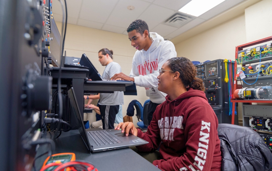 two students working on computers. one looks over at the other's screen. equipment and wires are in the background.