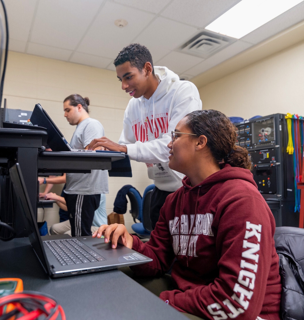 two students working on computers. one looks over at the other's screen. equipment and wires are in the background.