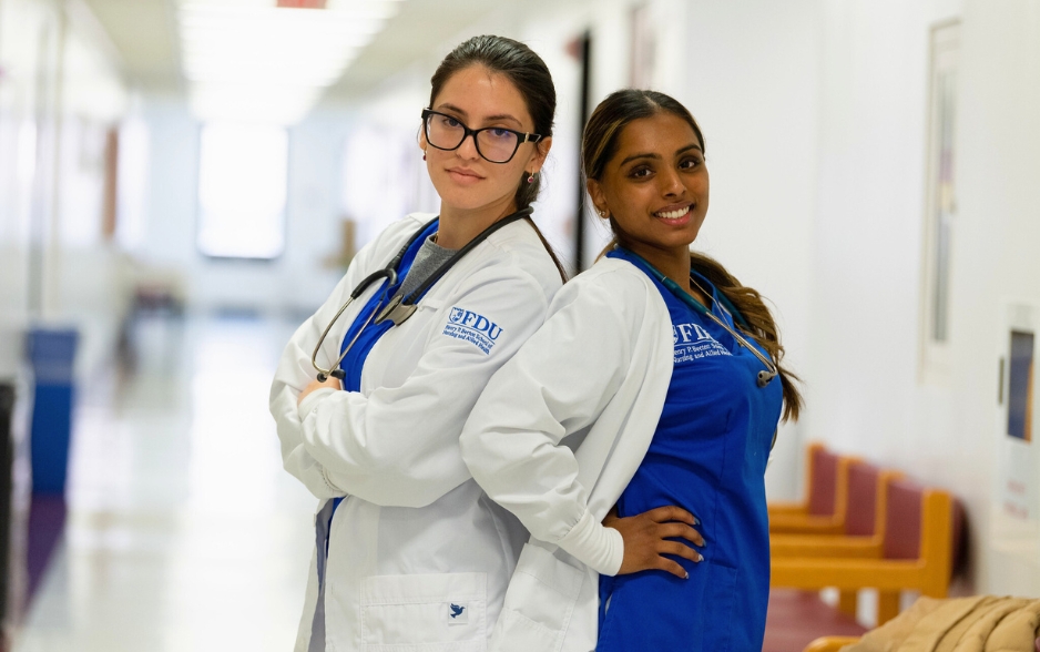 two students stand back to back. they both wear nurse uniforms and lab coats. one student crosses her arms and the other student has her hands on her hips.