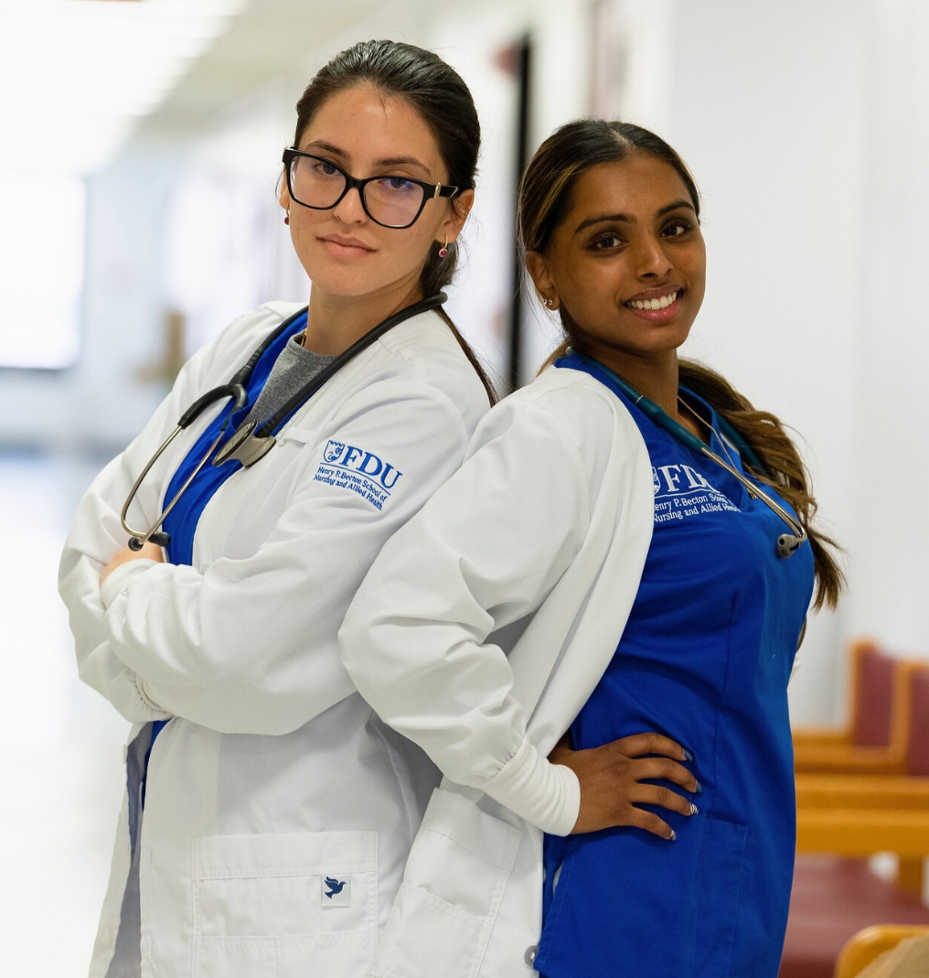 two students stand back to back. they both wear nurse uniforms and lab coats. one student crosses her arms and the other student has her hands on her hips.