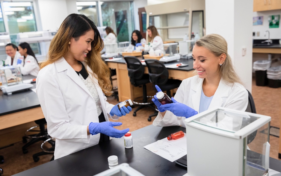 two students in lab coats work together. they are both wearing rubber gloves and hold pill bottles.