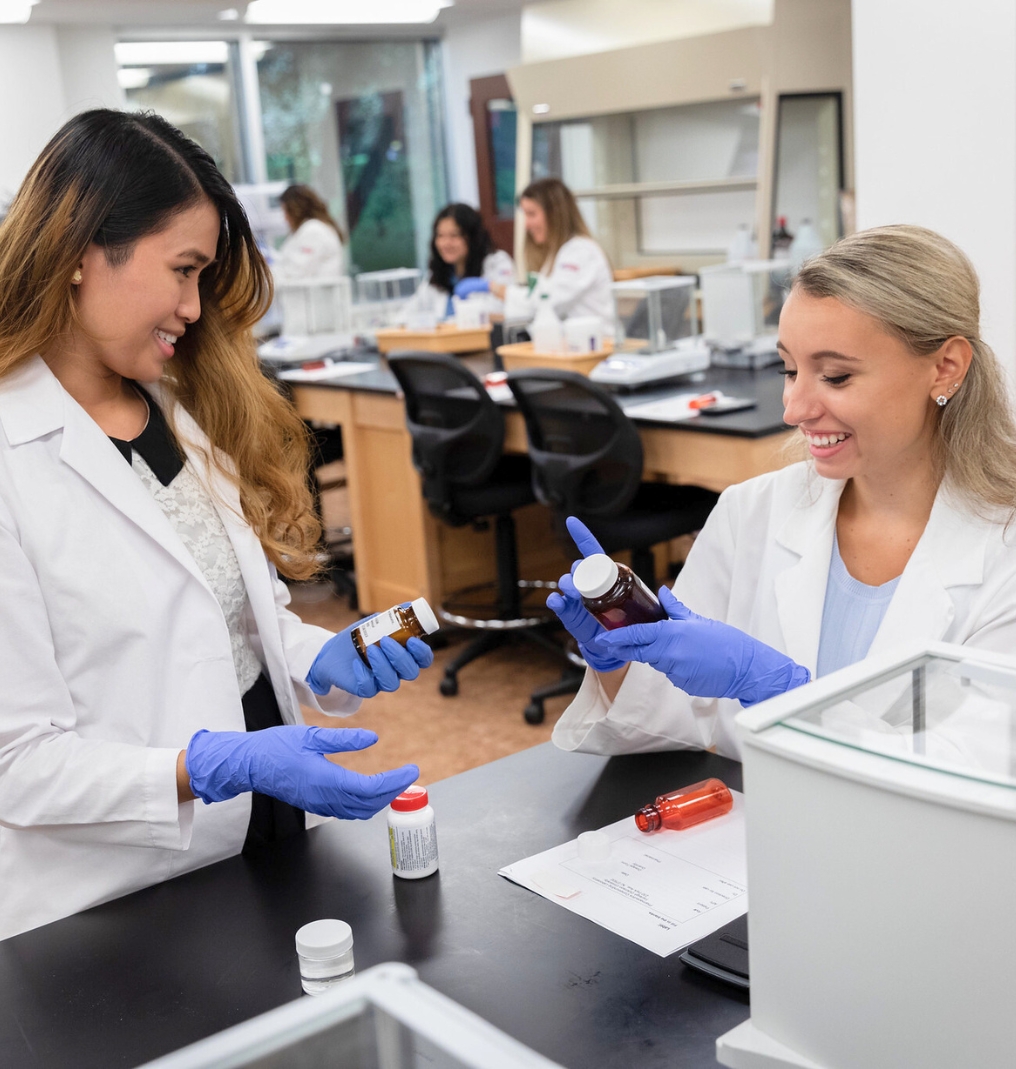 two students in lab coats work together. they are both wearing rubber gloves and hold pill bottles.