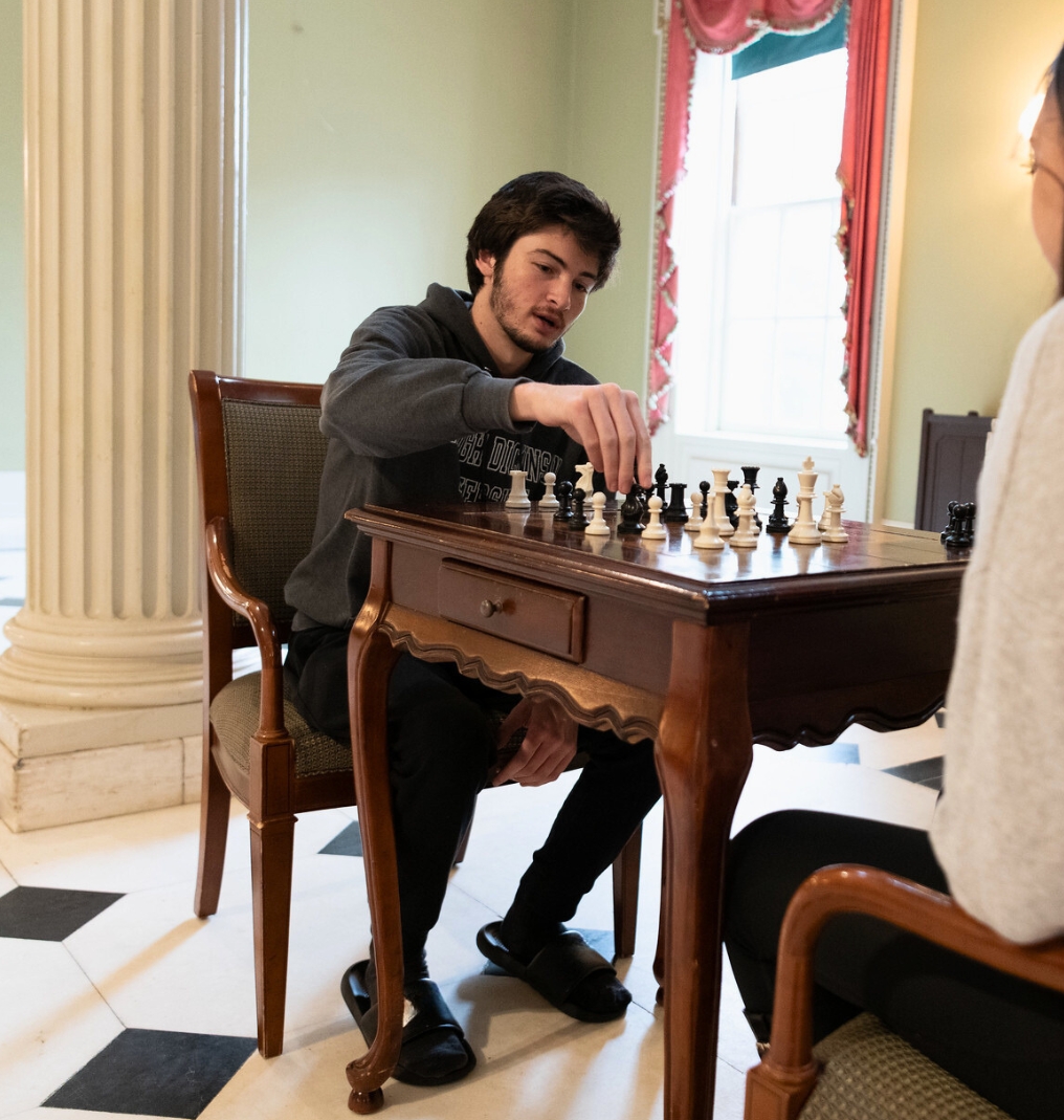 a student sits and plays chess. he wears an FDU hoodie. the back of another student playing against him can be seen in the edge of the photo.