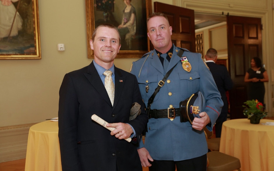 two men smile and pose for the camera. one wears a suit with an american flag pin. the other wears a police sergeant uniform.