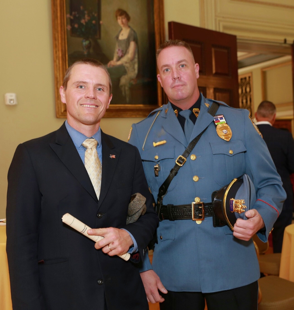 two men smile and pose for the camera. one wears a suit with an american flag pin. the other wears a police sergeant uniform.