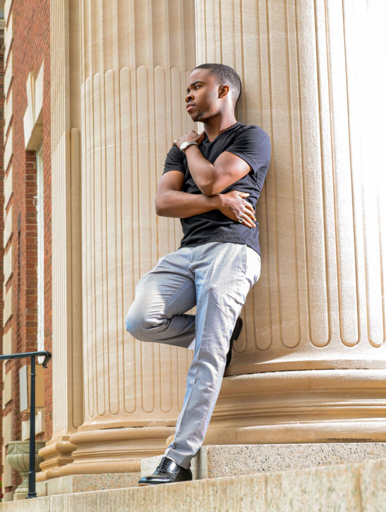 A young man leans back against the exterior pillar of a large brick building.