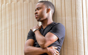 A young man leans back against the exterior pillar of a large brick building.