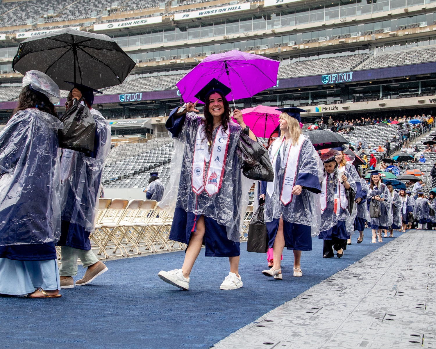 graduates walking down a pathway. they wear ponchos and hold umbrellas. one graduate smiles and holds up a peace sign hand signal.