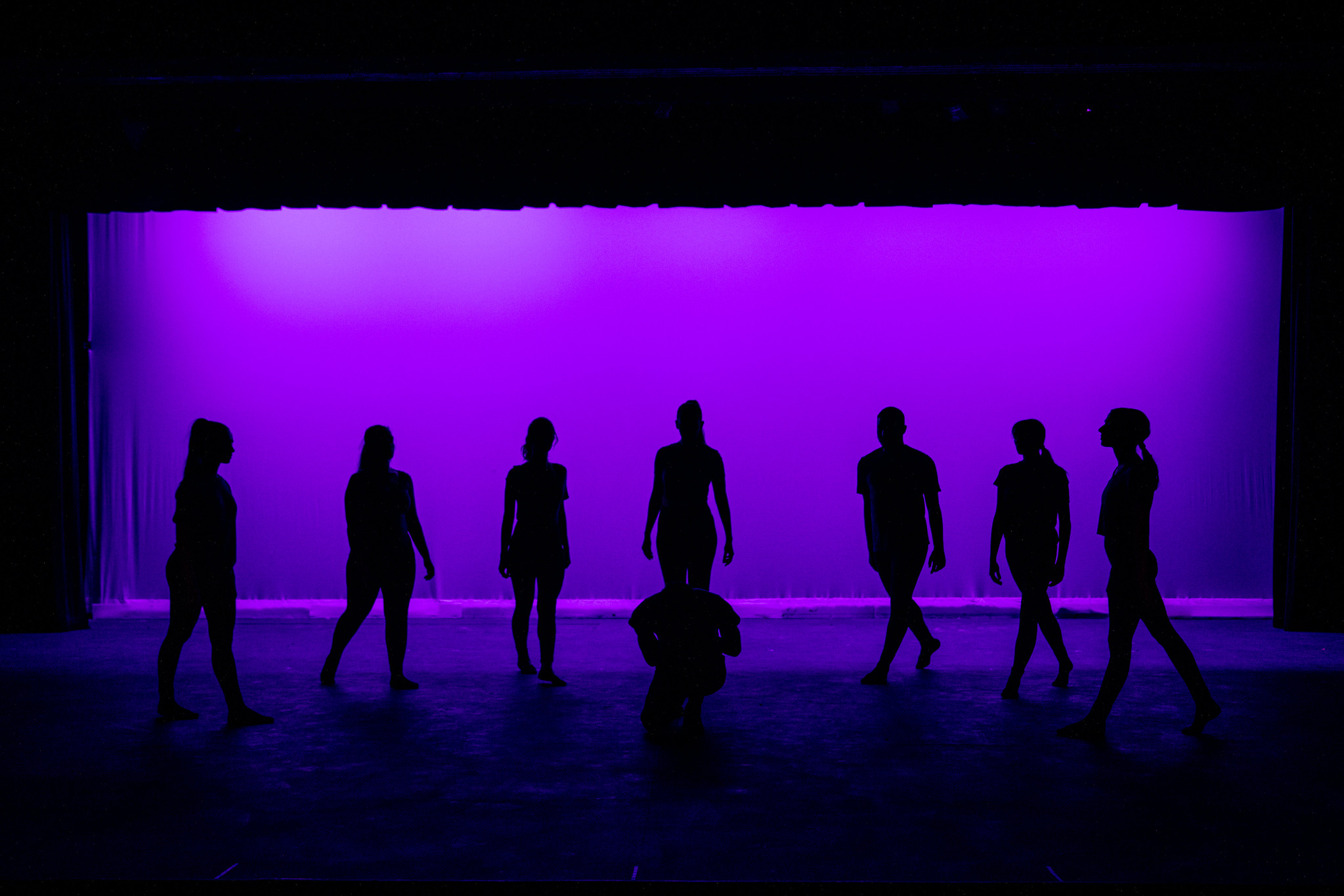 A dark stage is backlight with purple lights and dancers stand in shadow.