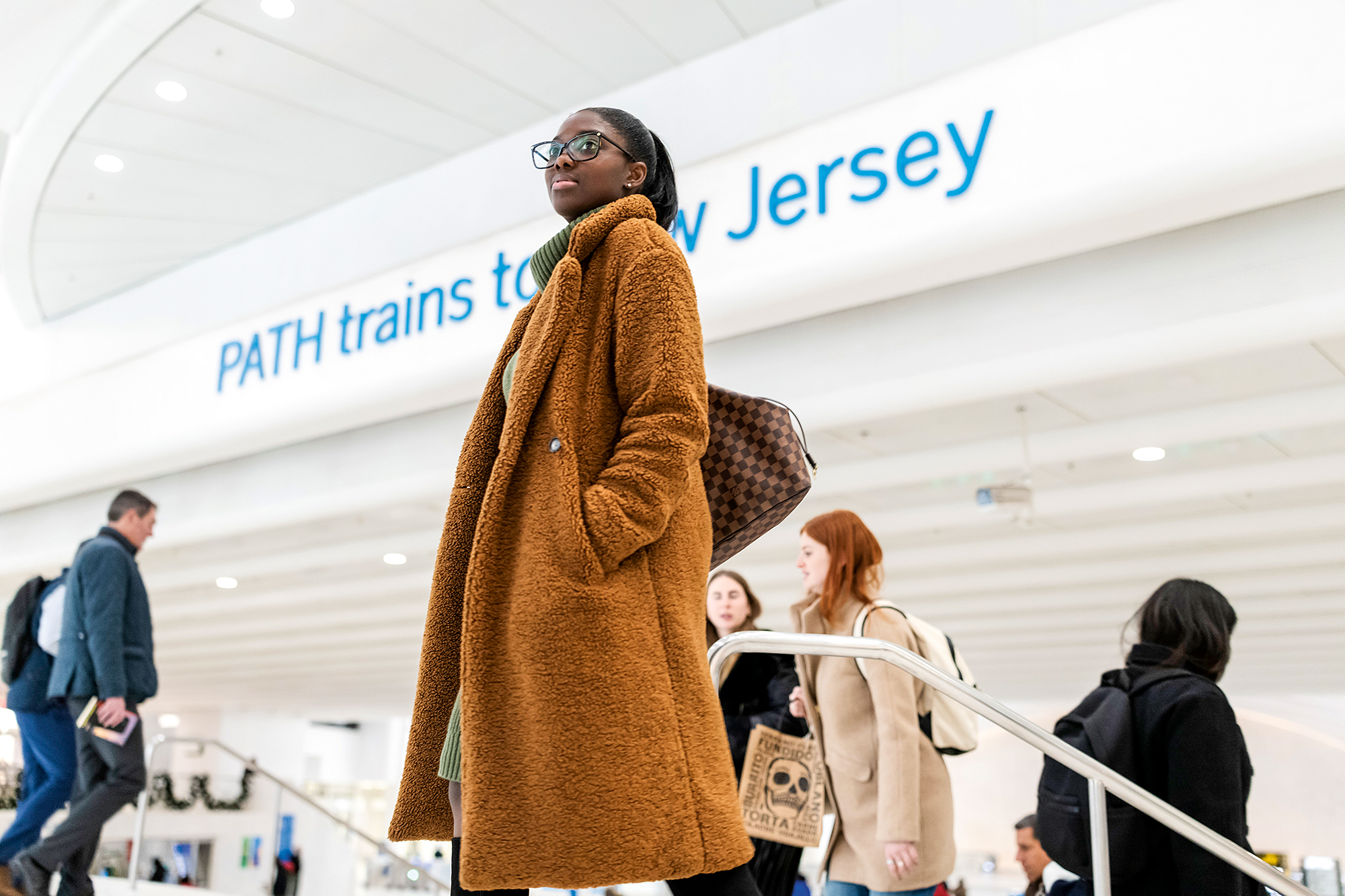 A woman wearing glasses and a winter coat pauses near the entrance to the NJ PATH trains in a train station.