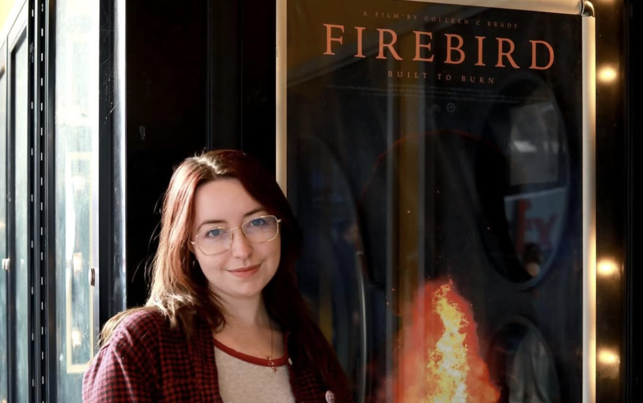 A young woman wearing glasses poses next to a documentary movie poster.