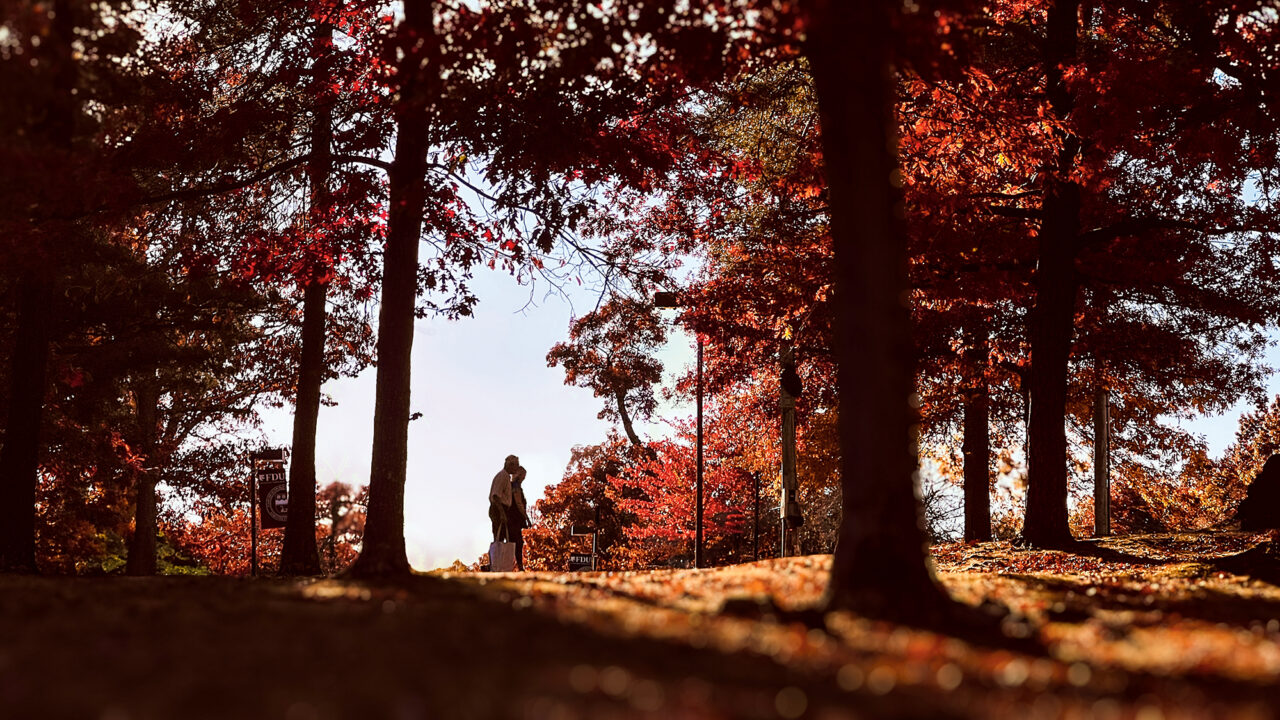 Two people, in shadows, take a stroll on the Florham Campus in fall, as sunlight streams through the orange leaves and trees.