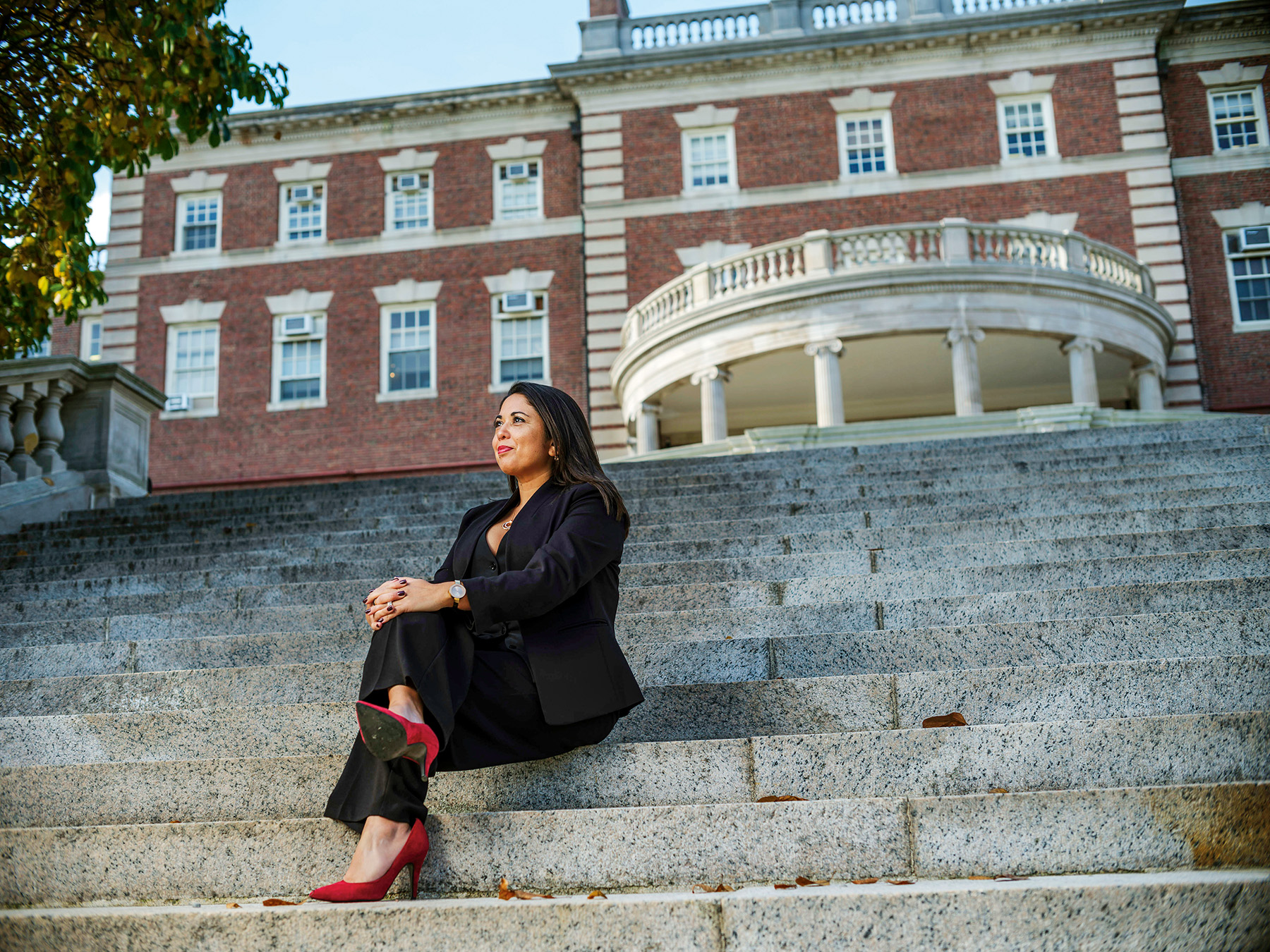 A woman wearing a suit and red high heels sits, legs crossed ona steep outdoor staircase. An imposing brick building is behind her.