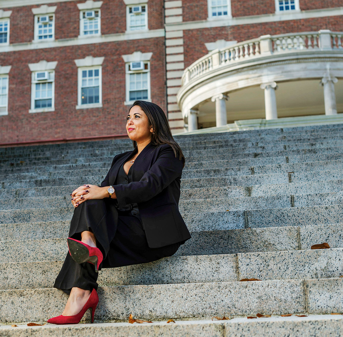 A woman wearing a suit and red high heels sits, legs crossed ona steep outdoor staircase. An imposing brick building is behind her.