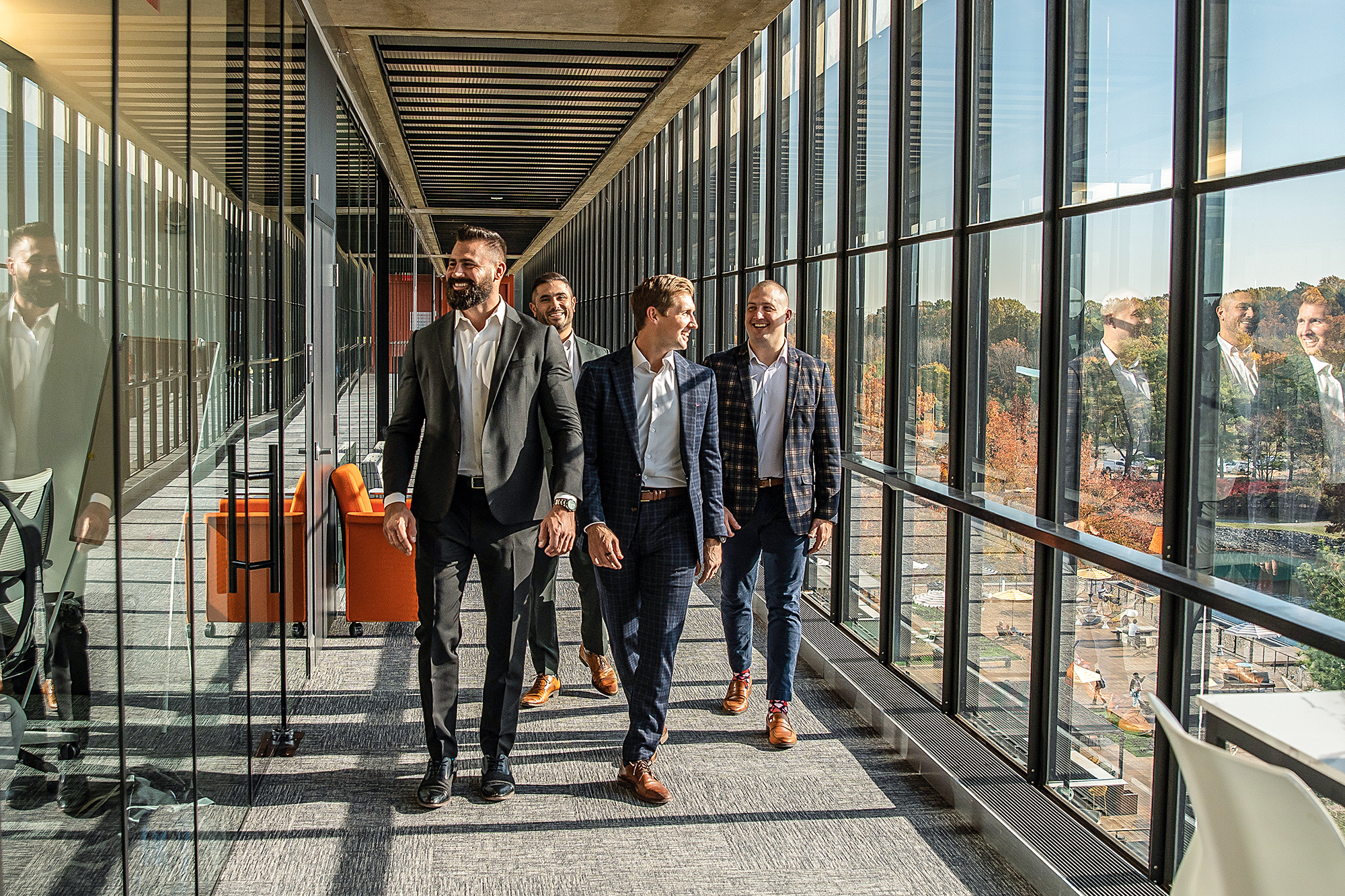 Four men wearing suits stride down a hall with a full wall of windows.