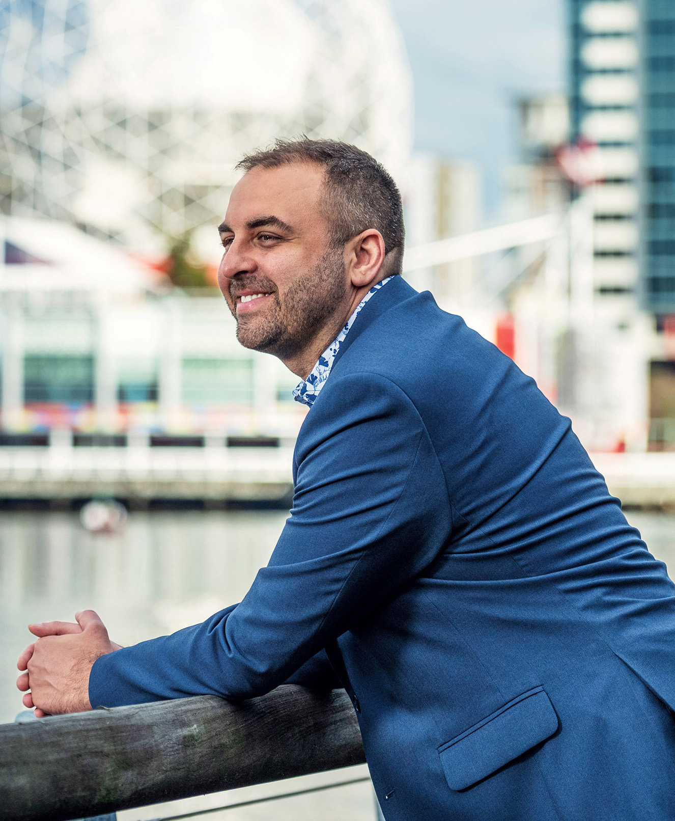 A man leans against a railing at the water's edge. He wears a blue suit and behind him, the Science World museum is visible in the Vancouver skyline.