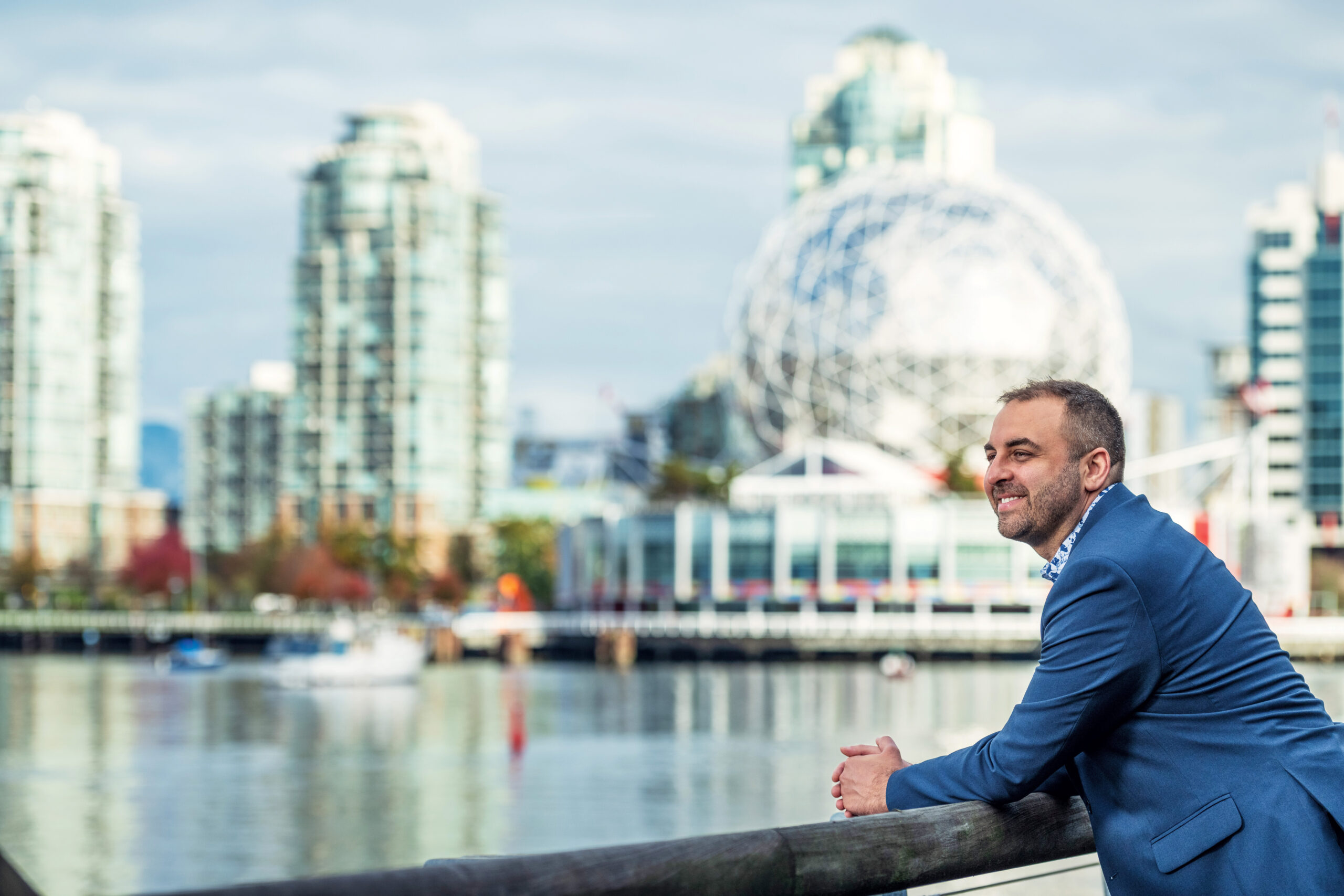 A man leans against a railing at the water's edge. He wears a blue suit and behind him, the Science World museum is visible in the Vancouver skyline.