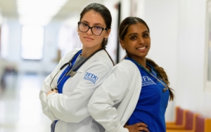 two students stand back to back. they both wear nurse uniforms and lab coats. one student crosses her arms and the other student has her hands on her hips.