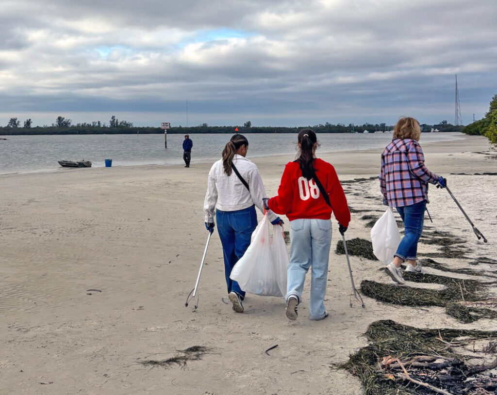 Three women walk on a beach carrying trash bags and collecting garbage.